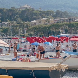 May include: A beach scene with people relaxing on the sand under red and blue umbrellas. There are boats on the shore and a white picket fence in the background.