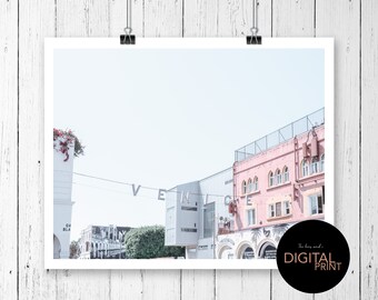 May include: A photograph of a pink building with the word "Venice" spelled out in white letters on a string hanging above the street. The building has a white fence and a white awning. The sky is a light blue.