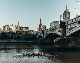 May include: A rowing team of four people in a boat on a river in front of a bridge and a city skyline. The bridge is made of stone and has arches. The city skyline is in the background and includes tall buildings and a church steeple.