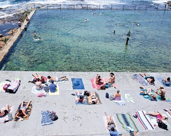 May include: A view from above of a public outdoor pool with people swimming and sunbathing on the surrounding deck. The pool is surrounded by a concrete deck with colorful towels and people relaxing on them.