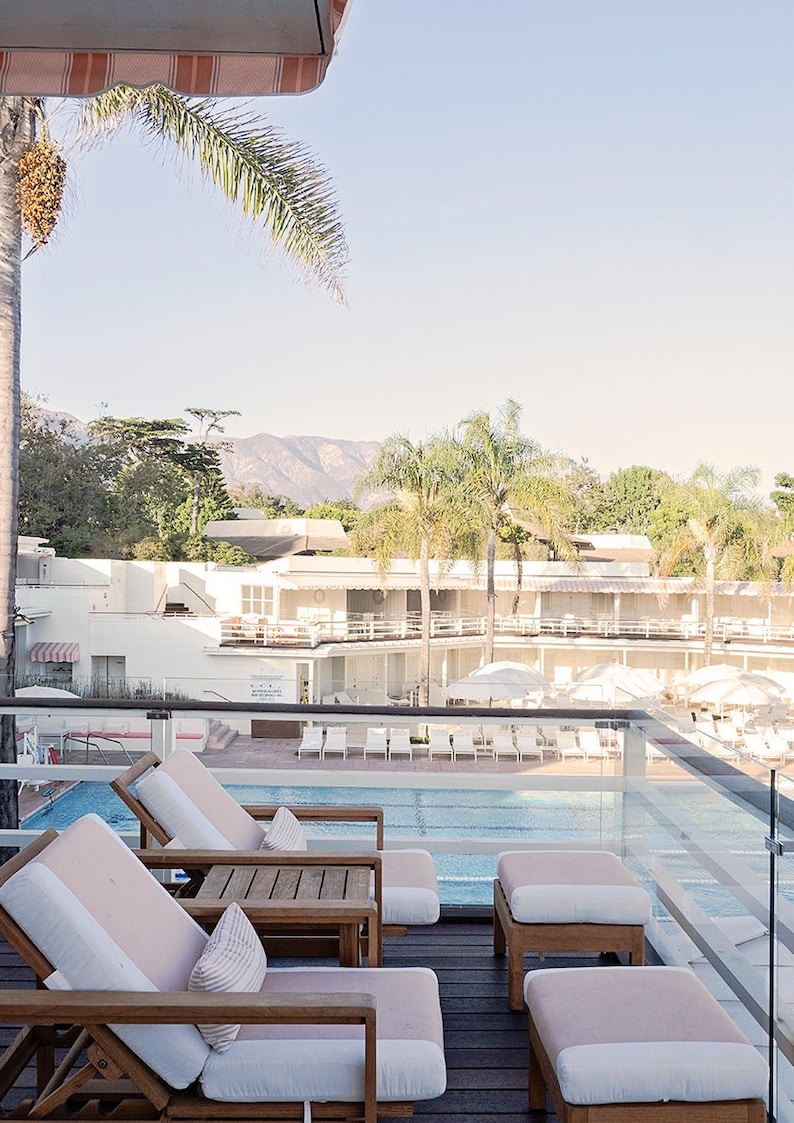 May include: A view from a balcony overlooking a swimming pool with lounge chairs and a mountain range in the background. There are three wooden chairs with pink cushions and a glass railing.