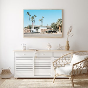May include: A white mid-century modern house with a black car parked in front of it. The house is surrounded by palm trees and a blue sky. The image is framed and hanging on a white wall above a white dresser with three drawers.