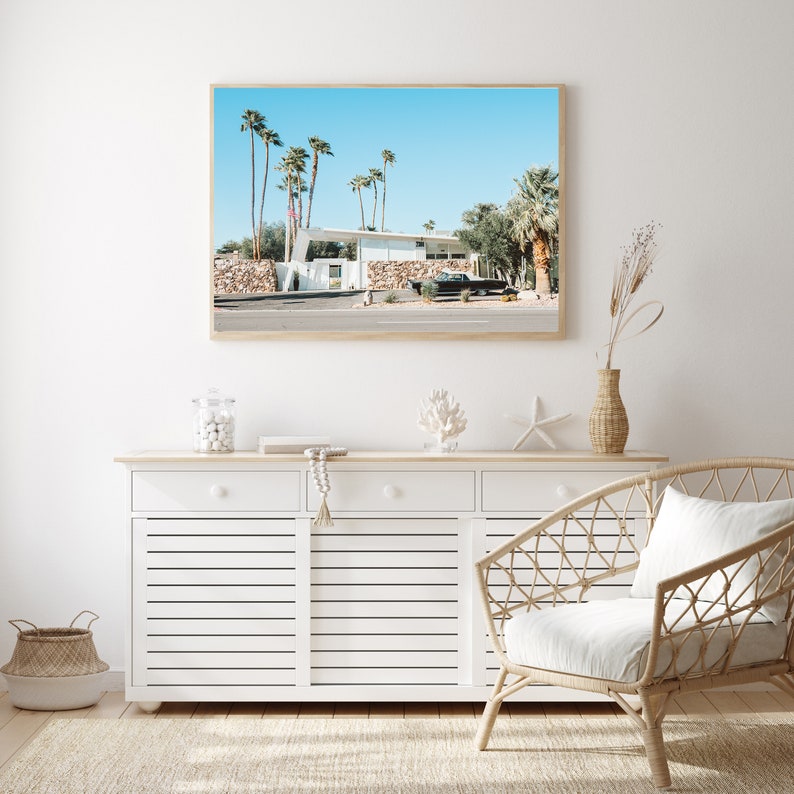 May include: A white mid-century modern house with a black car parked in front of it. The house is surrounded by palm trees and a blue sky. The image is framed and hanging on a white wall above a white dresser with three drawers.