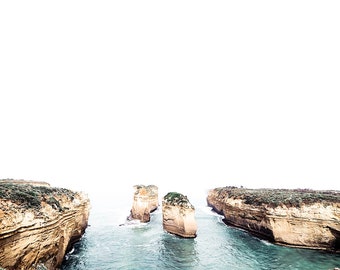 May include: A view of three rock formations jutting out of the ocean, with a narrow channel of water between them. The rocks are a light brown color and are surrounded by blue water. The water is calm and there is a slight mist in the air.