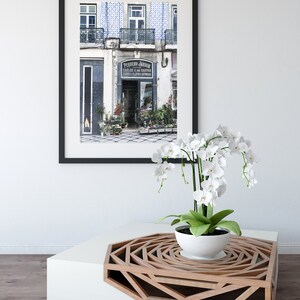 May include: A framed photograph of a flower shop in Lisbon, Portugal, with the text "PEQUENO JARDIM" above the entrance. The shop is decorated with blue and white tiles. A white orchid plant is sitting on a white and wood coffee table with a geometric design.