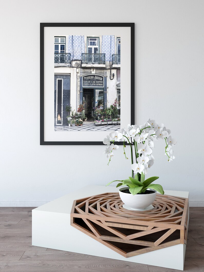 May include: A framed photograph of a flower shop in Lisbon, Portugal, with the text "PEQUENO JARDIM" above the entrance. The shop is decorated with blue and white tiles. A white orchid plant is sitting on a white and wood coffee table with a geometric design.