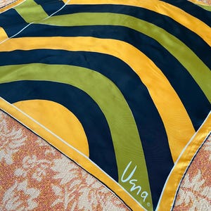 May include: A yellow, green, and black patterned silk scarf with a white border and the word "Usna" in white script in the bottom right corner.