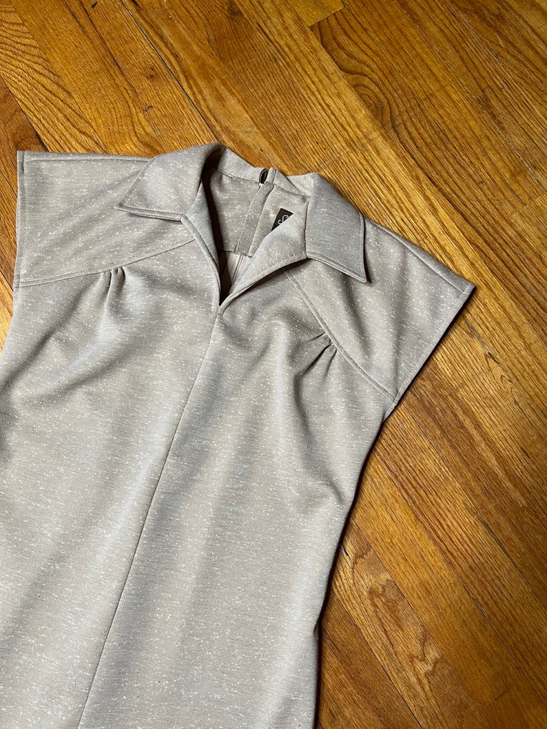 May include: A beige sleeveless dress with a collared neckline and a zipper closure in the back. The dress has a textured fabric with a subtle sparkle.