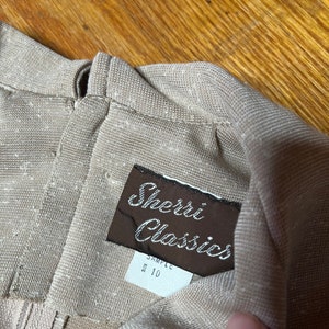 May include: A brown label with the text "Sherri Classics" and "Sample Size 10" attached to a beige fabric garment.