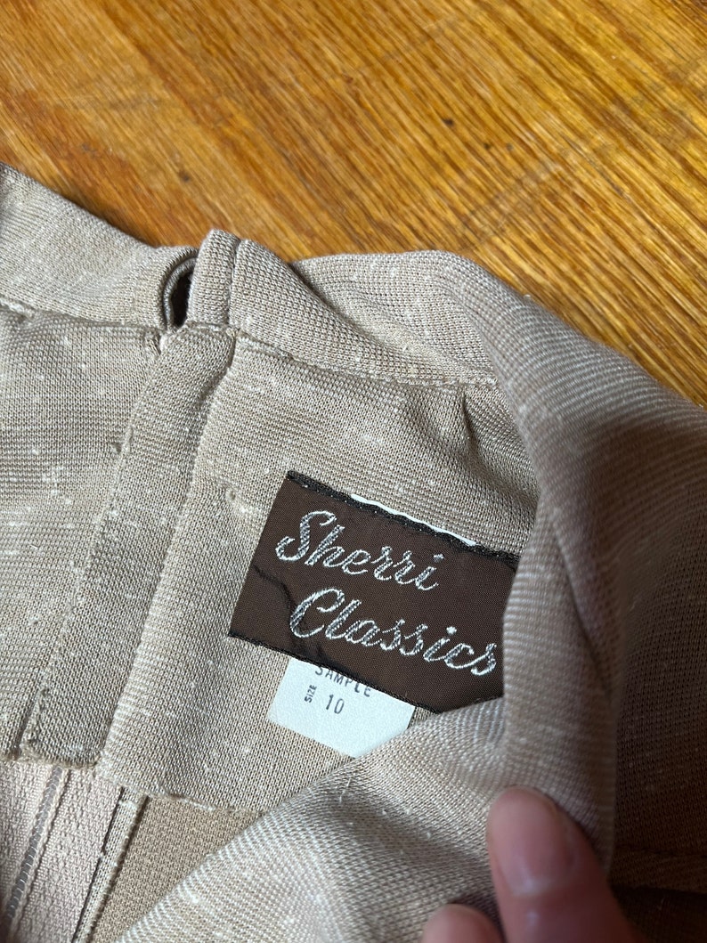 May include: A brown label with the text "Sherri Classics" and "Sample Size 10" attached to a beige fabric garment.