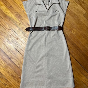 May include: A beige sleeveless dress with a collared neckline and a brown belt. The dress has a textured fabric and a straight silhouette.