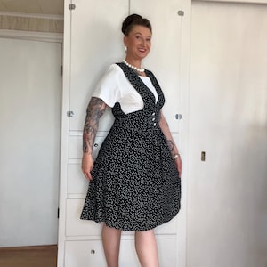 May include: A black and white polka dot dress with a white top underneath. The dress has a flared skirt and is knee-length. The person is wearing black and white shoes with a low heel. The background includes a white cabinet and a white door.