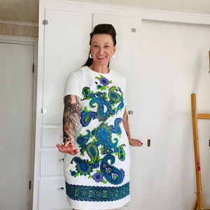 May include: A white short-sleeved shift dress featuring a vibrant paisley pattern in shades of blue, green, and purple. The design includes a prominent floral and teardrop motif across the front and a decorative geometric border along the hemline.