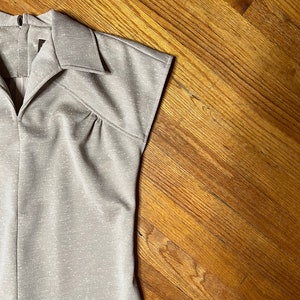 May include: A close-up of the top of a beige sleeveless dress with a collared neckline and a zipper closure. The dress has a subtle shimmer and is made of a textured fabric.