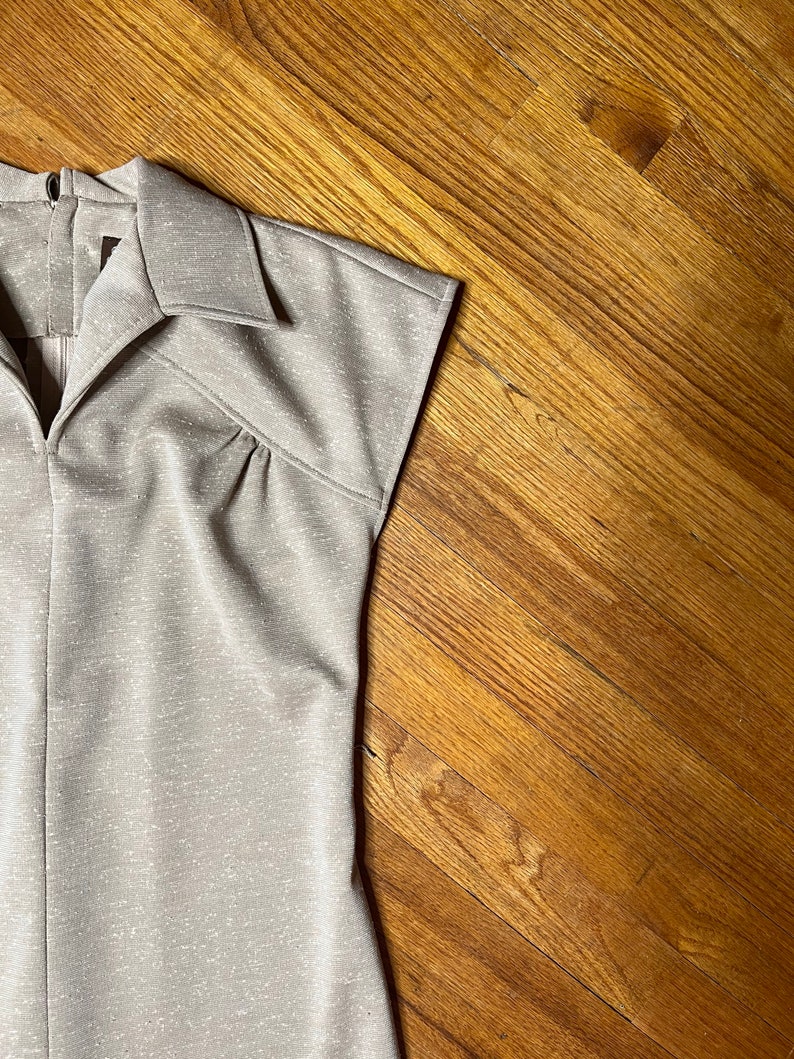 May include: A close-up of the top of a beige sleeveless dress with a collared neckline and a zipper closure. The dress has a subtle shimmer and is made of a textured fabric.