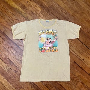 May include: A light yellow t-shirt with a cartoon character holding a beer mug and a bottle of beer. The text on the shirt reads "Beer Drinkers Make Better Lovers".