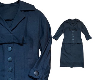May include: A black two-piece suit with a long-sleeved jacket and a skirt. The jacket has a collar, a button closure, and a bow detail at the waist. The skirt is knee-length and has a straight silhouette.
