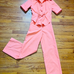 May include: A vintage coral two-piece outfit. The top has short sleeves, a collared neckline, and a button-down front. The waist is cinched with a drawstring. The matching trousers are wide-legged.