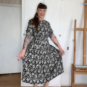 Może przedstawiać: Czarna sukienka w stylu vintage z kwiatowym nadrukiem, krótkimi rękawami i marszczoną talią. Sukienka ma biały kwiatowy wzór i jest połączona z brązowymi sandałami. Sukienka sięga do kolan i ma klasyczną, kobiecą sylwetkę.