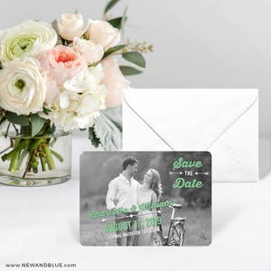 May include: Black and white photo save the date magnet with a couple in front of a bicycle. The text reads "Save the Date" in green with arrows on either side. The couple's names are "Charlotte & William" and the date is "August 17, 2019".