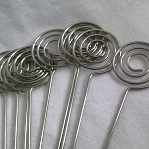May include: A collection of silver-colored spiral memo holders. Each holder features a circular spiral design at the top, connected to a thin, straight stem. The holders are arranged closely together, suggesting a set for displaying notes or photos.