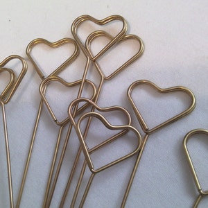 May include: A collection of gold-colored wire heart-shaped picks. The hearts are formed from a continuous wire, with a long, thin stem. The picks are arranged in a slightly overlapping pattern, suggesting a decorative or craft item.