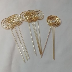 May include: A collection of gold-colored wire spiral place card holders. Each holder features a circular spiral design atop a slender, straight stem. The holders are arranged on a white surface, showcasing their elegant simplicity.