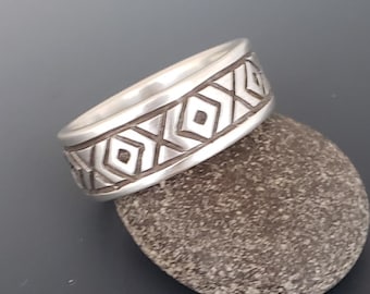 May include: A silver band ring featuring a repeating geometric pattern of etched diamonds and X-shaped lines. The metal has a polished finish, and the band has a consistent width throughout its circumference, showcasing a detailed engraved design.