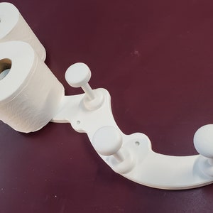 May include: White toilet paper holder with three hooks. The holder is designed to hold two rolls of toilet paper and has a curved shape. The hooks are round and are attached to the holder with a screw.