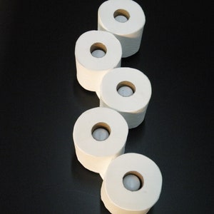 May include: Five white toilet paper rolls stacked on top of each other on a black surface.