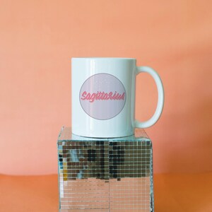 May include: White ceramic mug featuring the word "Sagittarius" in pink script within a pink circle, with a constellation design. The mug sits on a mirrored disco cube against a peach background.