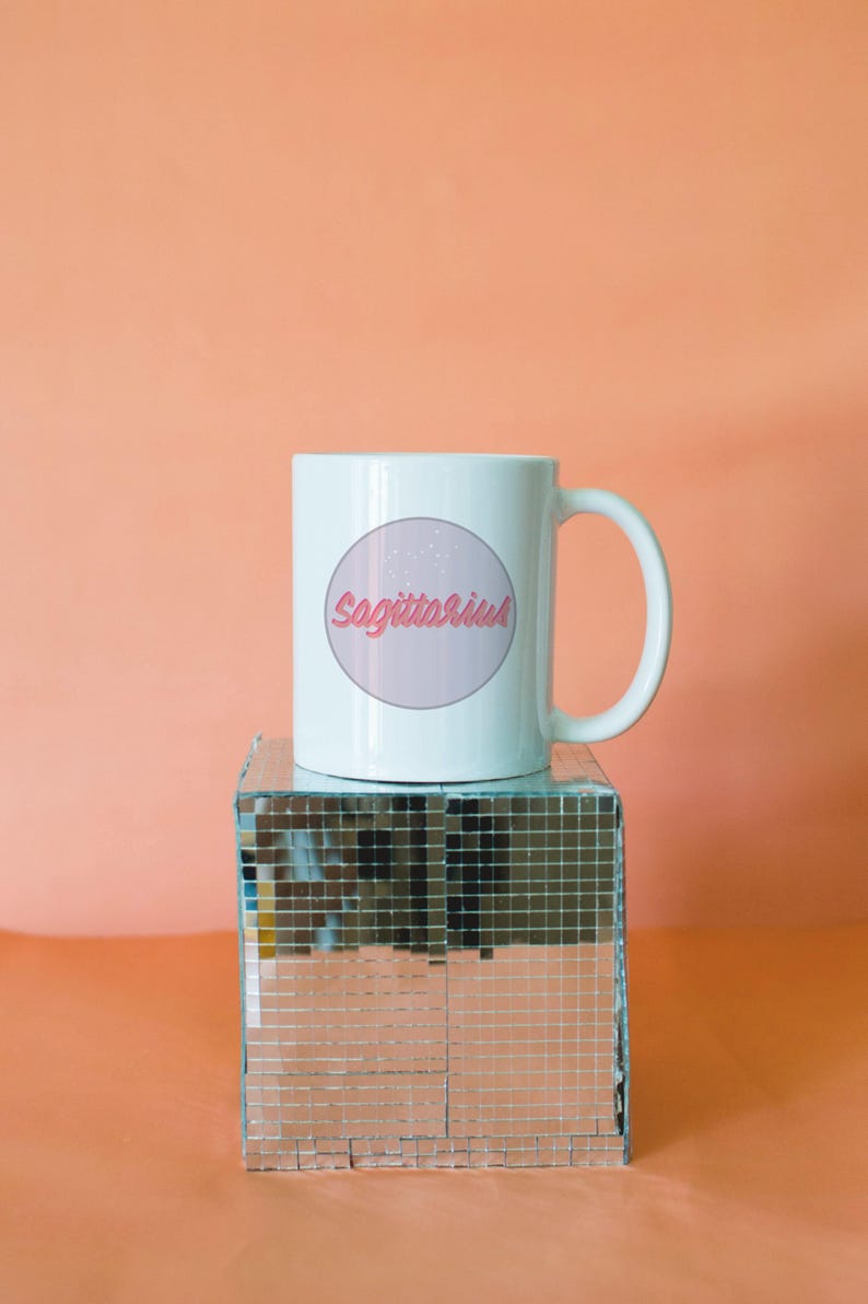 May include: White ceramic mug featuring the word "Sagittarius" in pink script within a pink circle, with a constellation design. The mug sits on a mirrored disco cube against a peach background.