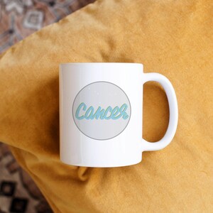 May include: White ceramic mug with the word "Cancer" in a light blue script inside a gray circle. The mug is on a yellow pillow.