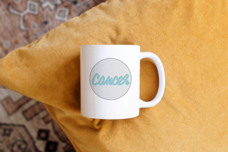 May include: White ceramic mug with the word "Cancer" in a light blue script inside a gray circle. The mug is on a yellow pillow.