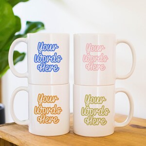 May include: Four white ceramic mugs stacked on top of each other. Each mug has a different coloured text that says "Your words here" in a cursive font. The mugs are on a wooden surface.