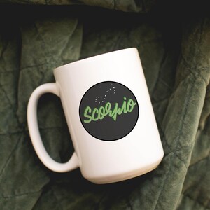May include: White ceramic mug with a black circle featuring the word "Scorpio" in green script, along with a constellation design. The mug is set against a dark green, textured background.