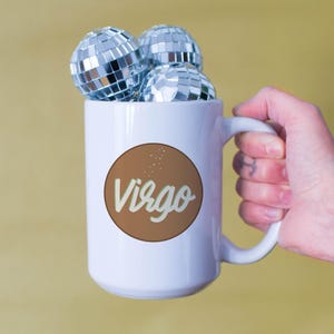 May include: White mug with a brown circle featuring the word "Virgo" in a script font. The mug is filled with three small disco balls. The mug is held by a hand against a yellow background.