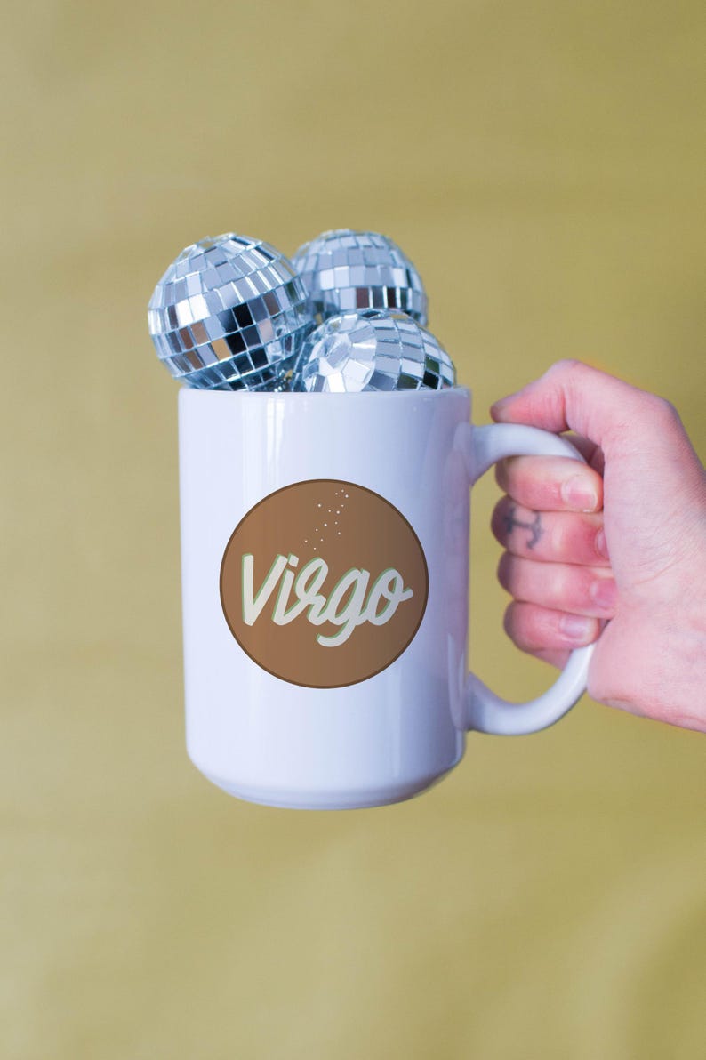 May include: White mug with a brown circle featuring the word "Virgo" in a script font. The mug is filled with three small disco balls. The mug is held by a hand against a yellow background.