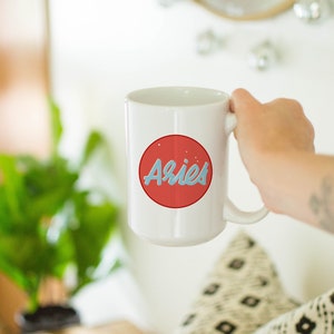 May include: White ceramic mug with a red circle design featuring the word "Aries" in light blue script. The mug is held by a person. The background includes a plant, a mirror, and a patterned pillow.