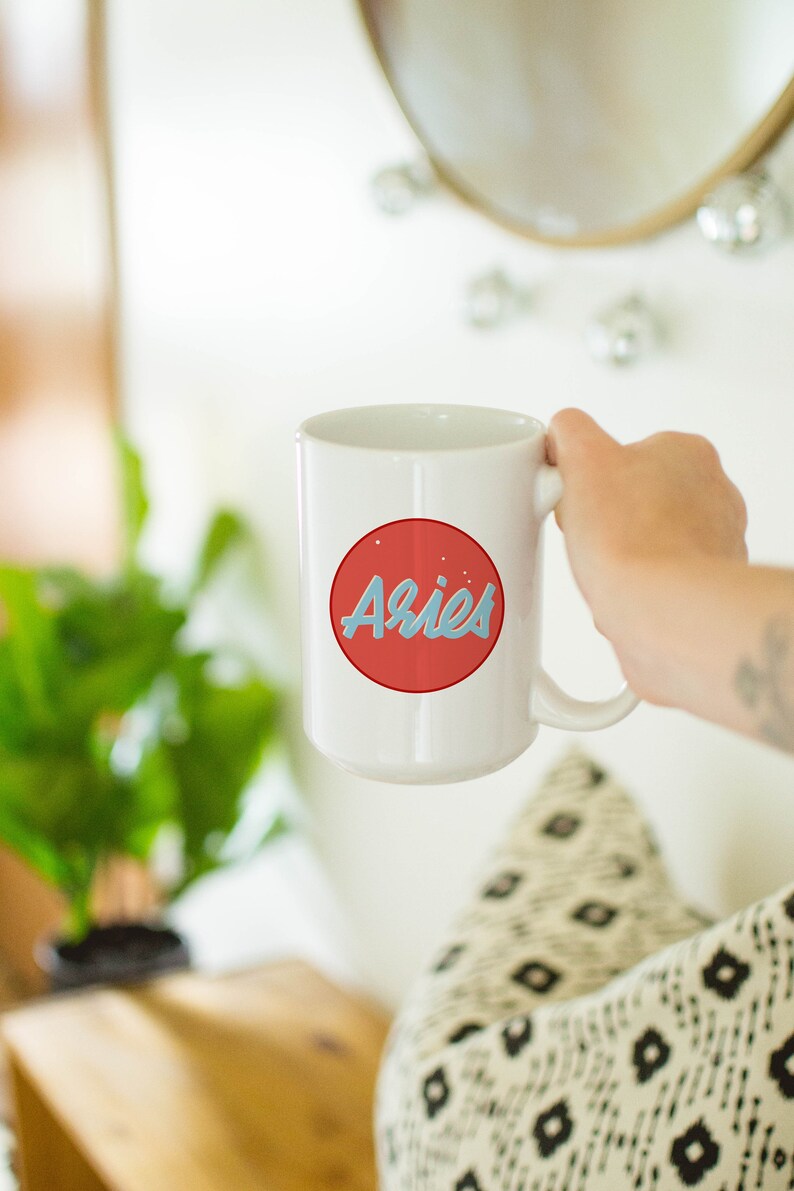 May include: White ceramic mug with a red circle design featuring the word "Aries" in light blue script. The mug is held by a person. The background includes a plant, a mirror, and a patterned pillow.