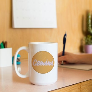 May include: White ceramic mug with a large handle, featuring a tan circle with the word "Gemini" in a light purple script. The mug is on a pink surface, with a person writing in the background. A calendar and pens are also visible.