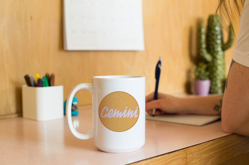 May include: White ceramic mug with a large handle, featuring a tan circle with the word "Gemini" in a light purple script. The mug is on a pink surface, with a person writing in the background. A calendar and pens are also visible.
