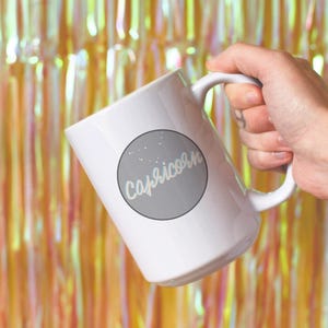 May include: White ceramic mug featuring the word "Capricorn" in a light blue script within a gray circle, with small white star accents. The mug is held against a backdrop of gold and pink shimmering tinsel.