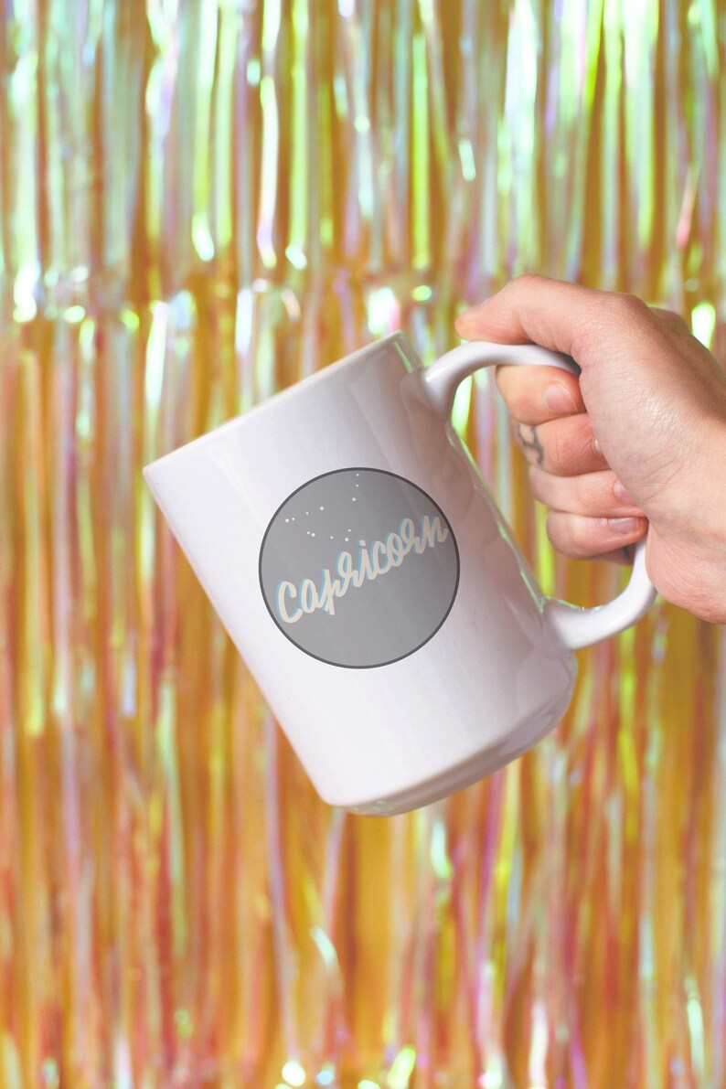 May include: White ceramic mug featuring the word "Capricorn" in a light blue script within a gray circle, with small white star accents. The mug is held against a backdrop of gold and pink shimmering tinsel.