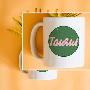 May include: White ceramic mug featuring the word "Taurus" in pink script within a green circle, with small white dots. The mug has a white handle and is set against an orange background with gold decorative elements.