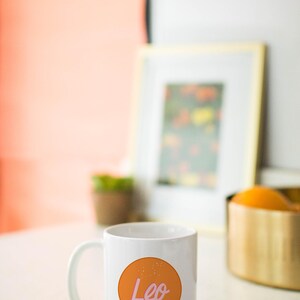 May include: White ceramic mug with a handle, featuring an orange circle with the word "Leo" in pink script. The mug is on a white surface, with a blurred background including a framed picture and a gold-colored container.