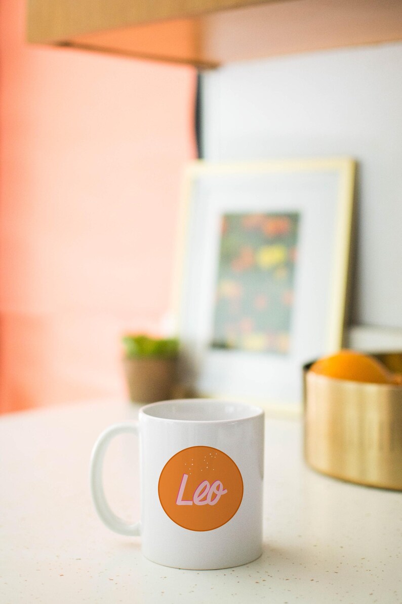 May include: White ceramic mug with a handle, featuring an orange circle with the word "Leo" in pink script. The mug is on a white surface, with a blurred background including a framed picture and a gold-colored container.