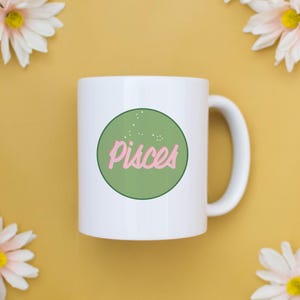 May include: White ceramic mug with a green circle containing the word "Pisces" in pink script. The mug is surrounded by white and yellow daisies on a yellow background.