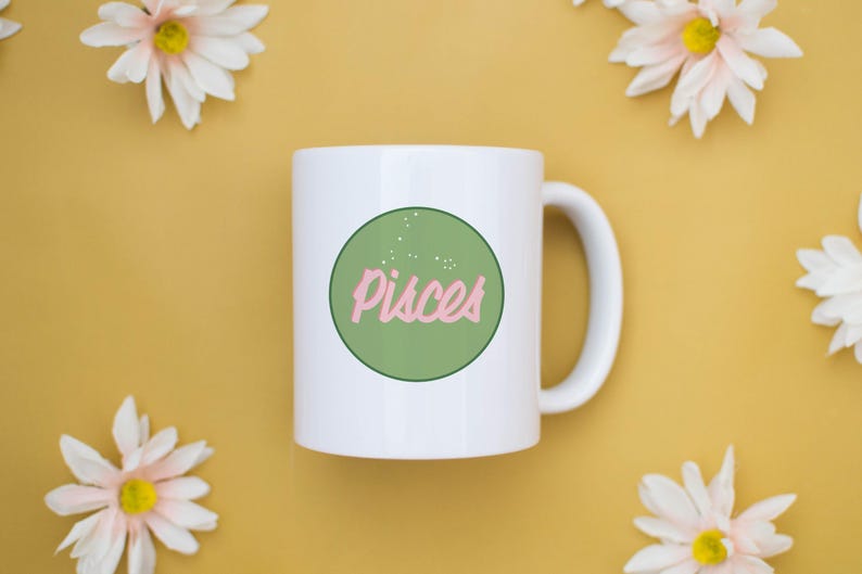 May include: White ceramic mug with a green circle containing the word "Pisces" in pink script. The mug is surrounded by white and yellow daisies on a yellow background.
