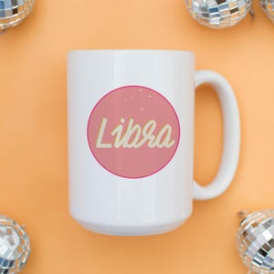 May include: White ceramic mug with a pink circle containing the word "Libra" in a cursive font. The mug is on an orange surface, with disco ball ornaments in the background.